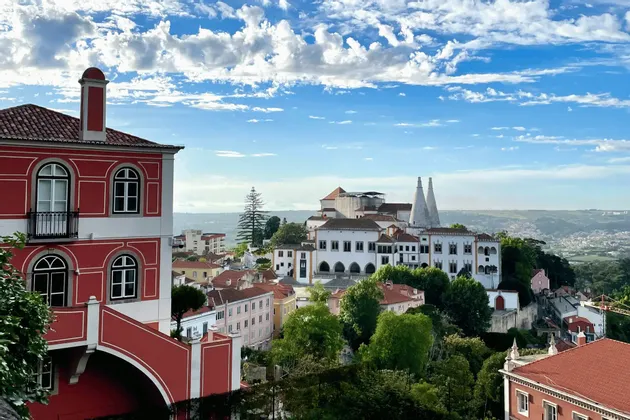A red and white building overlooks a historic town with a palace featuring two white conical chimneys, under a partly cloudy sky.