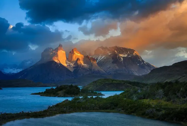 Cime montuose frastagliate illuminate dalla luce dorata del sole sotto un cielo scuro e nuvoloso, con un lago blu e calmo in primo piano.