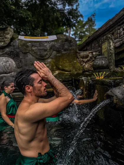 Ein Mann von einer WeRoad Gruppenreise nimmt an einem Wasserreinigungsritual teil, betend mit zusammengelegten Händen unter einem Steinbrunnen.