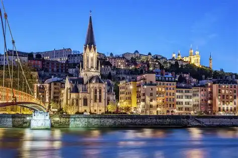 Eine Stadtlandschaft in der Dämmerung mit einer beleuchteten Kirche und Gebäuden entlang eines Flusses, wobei sich Lichter auf dem Wasser spiegeln.
