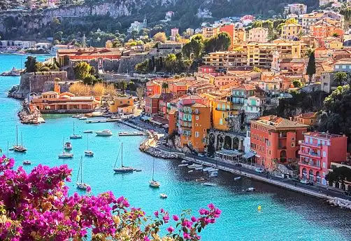 An aerial view of a colorful coastal town with boats docked in a turquoise bay, framed by bright pink flowers.