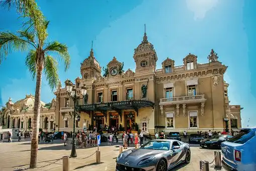 A grand, ornate building with a plaza full of people and luxury cars on a sunny day.