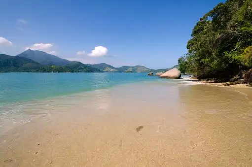 A sandy beach with clear, shallow water leading to a bay with green mountains in the distance under a blue sky.