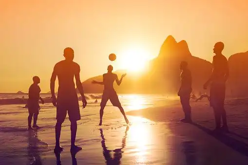 A WeRoad group trip playing with a ball on a beach, silhouetted against a golden sunset with mountains in the background.