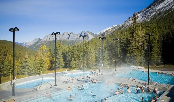 Persone che si rilassano in diverse piscine fumanti all'aperto, circondate da una fitta pineta con grandi montagne rocciose sullo sfondo.