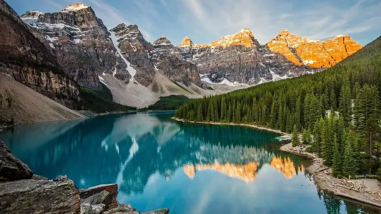 Montagne illuminate dal sole e coperte di neve che si riflettono in un lago calmo e turchese, circondato da una fitta pineta.