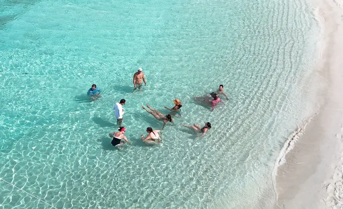 Eine Luftaufnahme einer WeRoad Gruppenreise, die in klarem, flachem türkisfarbenem Wasser an einem weißen Sandstrand schwimmt und entspannt.