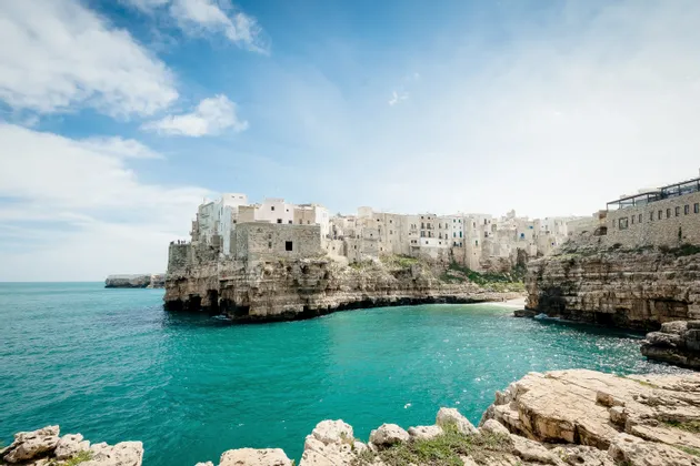 Un borgo con edifici bianchi e in pietra si erge su una scogliera rocciosa a picco su una baia turchese sotto un cielo azzurro parzialmente nuvoloso.