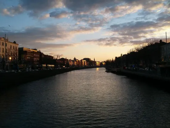 Un ampio fiume scorre attraverso una città al tramonto, con edifici che costeggiano le sponde sotto un cielo nuvoloso.