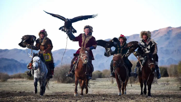 Quatre chasseurs à l'aigle en tenue traditionnelle à cheval, chacun tenant un aigle royal, dans un vaste paysage avec des montagnes en arrière-plan.
