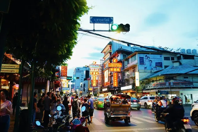 Una concurrida calle de la ciudad al anochecer con un tuk-tuk en un paso de cebra, tráfico, peatones y edificios con letreros de neón brillantes.