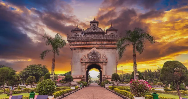Un grande e ornato monumento ad arco si erge in un parco pubblico, circondato da palme, sotto un drammatico cielo al tramonto.