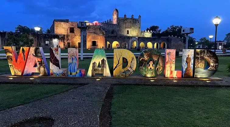 Lettere illuminate e colorate che compongono 'VALLADOLID' su un prato verde, con un vecchio edificio in pietra illuminato sullo sfondo al tramonto.