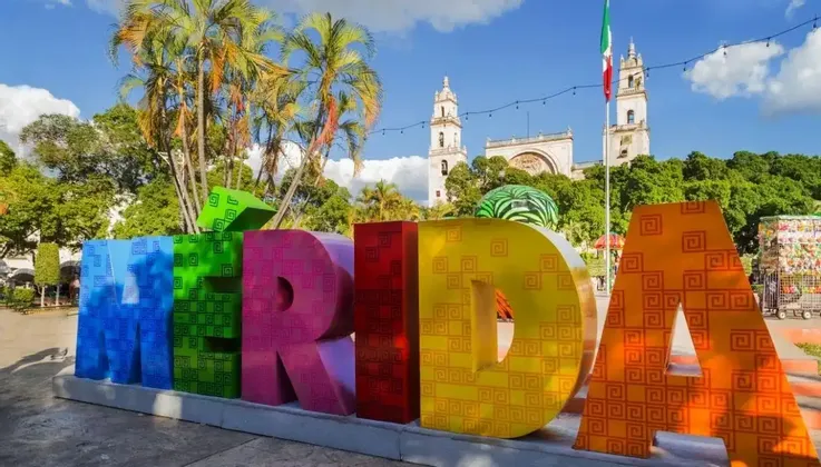 Grandi lettere colorate che compongono la parola 'MERIDA' si ergono in una piazza con una cattedrale, palme e una bandiera messicana sullo sfondo.