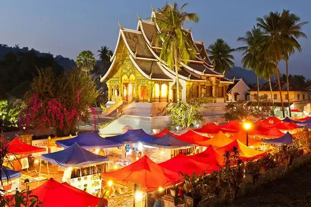Tende colorate rosse e blu di un mercato notturno brillano davanti a un tempio dorato illuminato al tramonto.