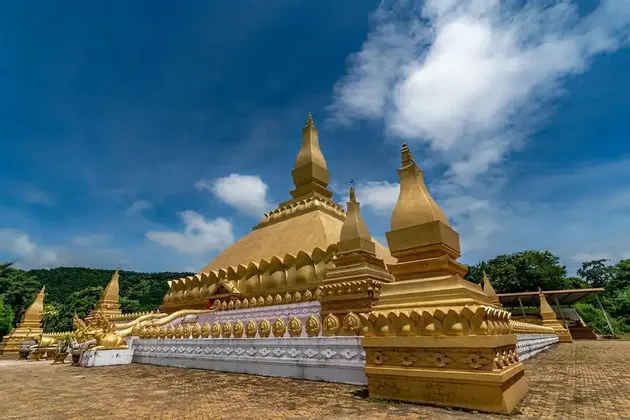 Una grande stupa dorata e riccamente decorata, con molteplici guglie, si erge in un cortile pavimentato sotto un cielo azzurro con nuvole.