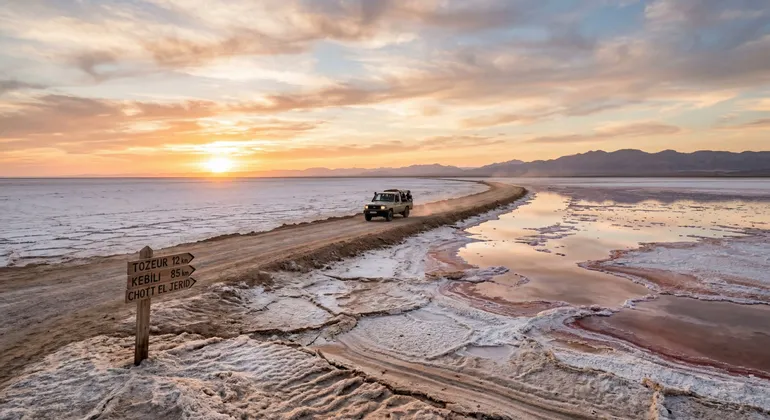 Ein Geländewagen einer WeRoad-Gruppenreise fährt bei Sonnenuntergang auf einem unbefestigten Damm über die Salzwüste Chott el Jerid.