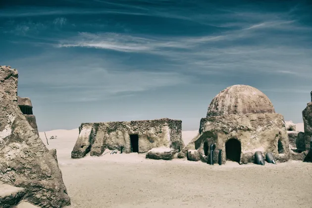 Sandfarbene Gebäude mit Kuppeldach stehen in einer weiten Wüste unter einem gestreiften blauen Himmel.
