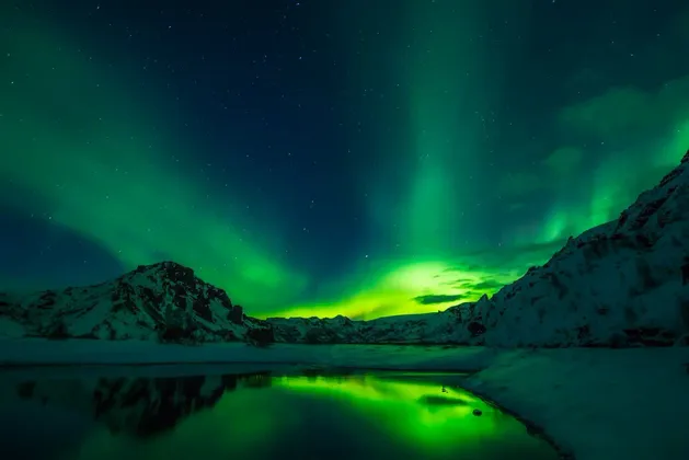 L'aurora boreale verde illumina un cielo notturno stellato sopra montagne innevate, riflettendosi in un lago calmo.