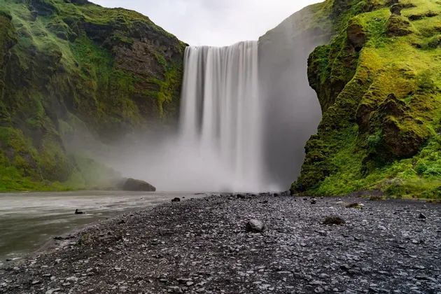 Uno scatto a lunga esposizione di una vasta cascata che si getta tra scogliere verdi e muschiose su una riva scura e rocciosa.