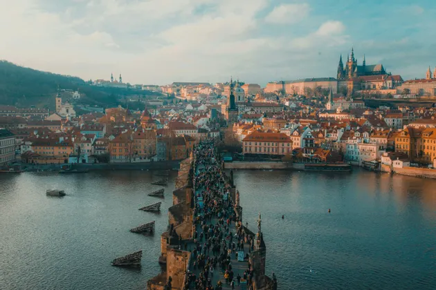 Una vista dall'alto di un ponte di pietra affollato su un fiume, con un panorama urbano europeo e un castello sullo sfondo.