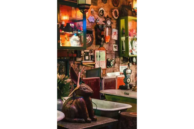 Una grande statua di coniglio si trova su un tavolo in una stanza piena di oggetti d'antiquariato, curiosità e vecchie vasche da bagno, contro un muro di pietra ingombro.