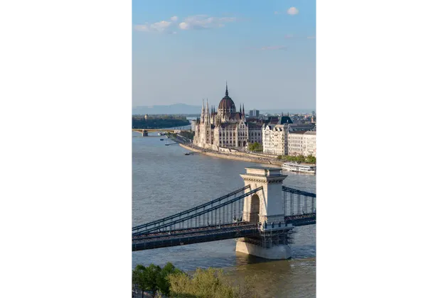 Un ponte a catena storico attraversa un ampio fiume, con un palazzo del parlamento ornato e a cupola sulla riva opposta sotto un cielo blu.