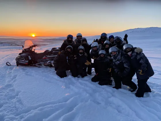 Un viaje en grupo de WeRoad posando para una foto en un paisaje nevado junto a una moto de nieve durante el atardecer.