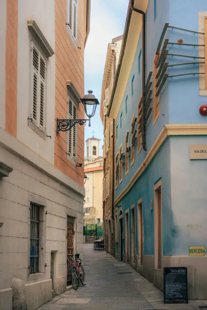 Un vicolo stretto acciottolato tra edifici colorati, con una bicicletta rossa parcheggiata e un campanile visibile in lontananza.