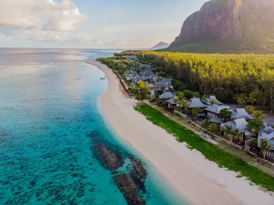 Una vista aerea di un resort su una spiaggia di sabbia bianca accanto ad acque turchesi, con una foresta lussureggiante e un'imponente montagna dietro.