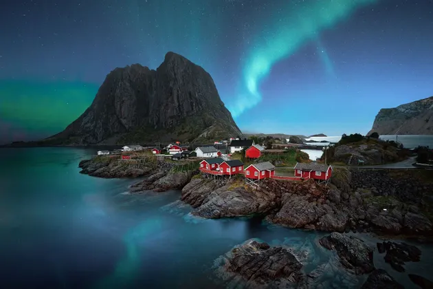 Cabine rosse su una costa rocciosa si ergono sotto una montagna, mentre l'aurora boreale verde risplende nel cielo notturno stellato.