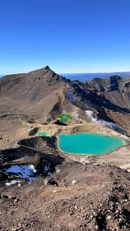 A high-angle view of three turquoise and green geothermal lakes steaming in a rocky, volcanic crater under a clear blue sky.