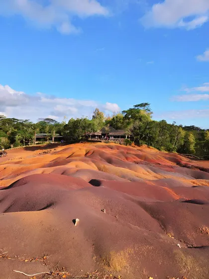 Un paesaggio di dune ondulate e multicolori nei toni del rosso e del viola, con una piattaforma panoramica e una foresta sullo sfondo.