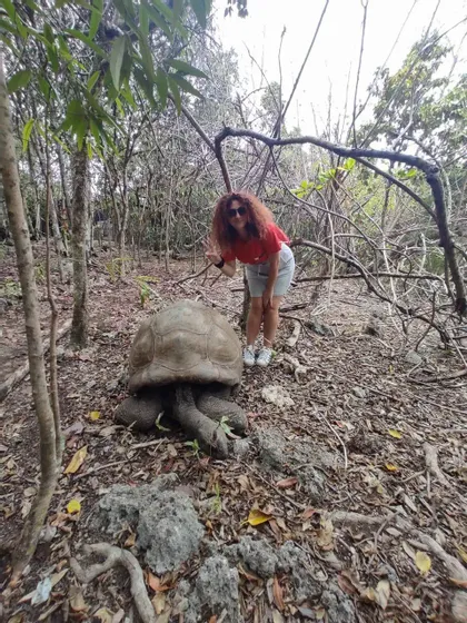 Una donna dai capelli rossi e ricci posa con il segno della pace accanto a una tartaruga gigante su un terreno foglioso in una foresta.