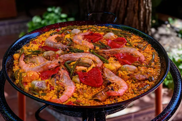 Un primo piano di una paella tradizionale in una grande padella nera, ricca di gamberi, peperoni rossi e fagiolini.
