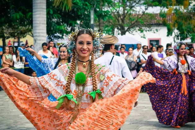 Eine Frau in einem farbenfrohen traditionellen Kleid und Kopfschmuck lächelt, während sie bei einer Freiluftparade tanzt.