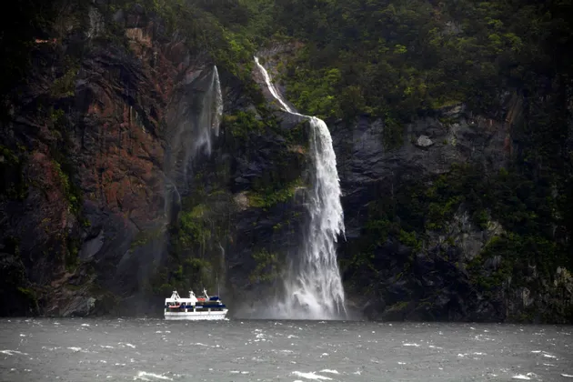 Un'imbarcazione turistica naviga in acque agitate ai piedi di due potenti cascate che si riversano da una scogliera rocciosa e lussureggiante.