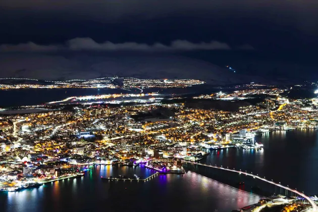 Veduta aerea di una città costiera innevata illuminata di notte, con un ponte illuminato che attraversa l'acqua e montagne sullo sfondo.