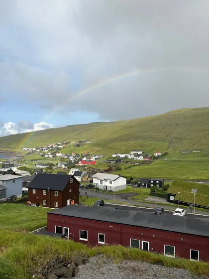 Un arcobaleno si inarca su un piccolo villaggio adagiato ai piedi di una grande collina verde sotto un cielo nuvoloso.