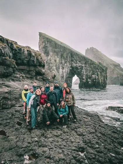Un gruppo WeRoad in posa su una costa rocciosa, con grandi faraglioni e un arco naturale alle spalle, sotto un cielo nuvoloso.