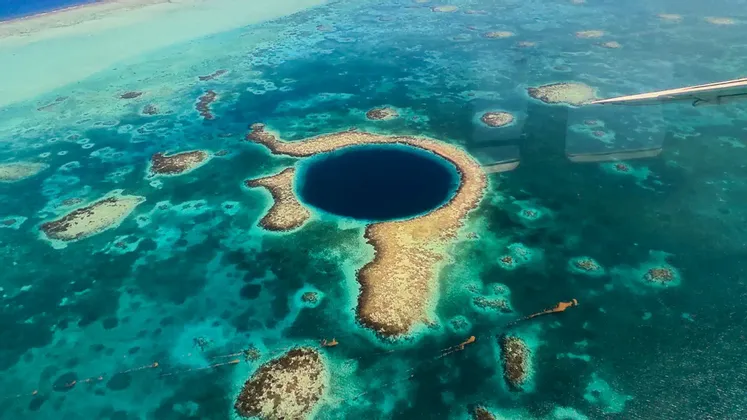 Veduta aerea di un grande buco blu circolare circondato da una barriera corallina in acque oceaniche turchesi, con l'ala di un aereo visibile.