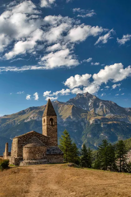 Una rustica chiesa in pietra con campanile si erge su una collina, con una vasta catena montuosa sullo sfondo, sotto un cielo azzurro e nuvoloso.