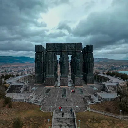 Veduta aerea di un imponente monumento con alte colonne scolpite, mentre persone camminano sulla sua piazza di pietra sotto un cielo nuvoloso.
