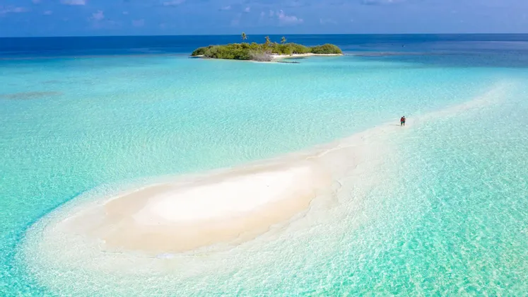 Veduta aerea di due persone su un banco di sabbia bianca che serpeggia in acque turchesi verso una piccola isola tropicale.