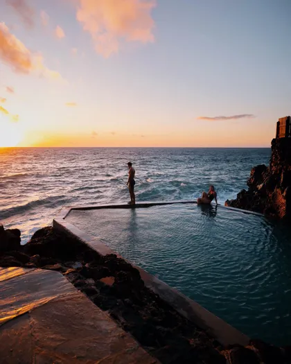 Un uomo in piedi e una donna seduta a bordo di una piscina a sfioro con vista sull'oceano, durante un tramonto colorato.