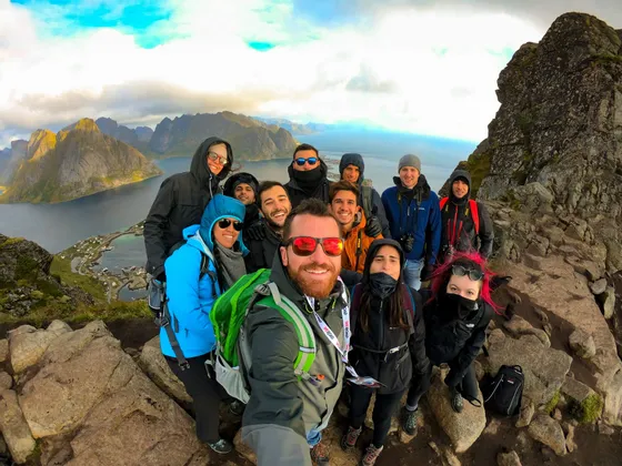 Eine WeRoad-Gruppenreise macht ein Selfie auf einem felsigen Berggipfel, mit Blick auf ein Küstendorf und Berge über eine Bucht.