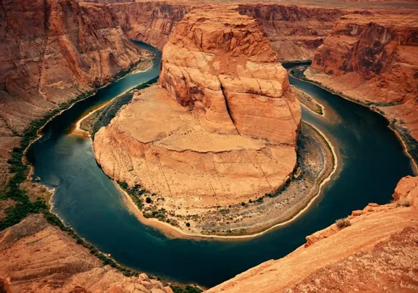 Un fiume blu scuro scorre in una curva a ferro di cavallo attorno a una grande formazione rocciosa centrale, all'interno di un profondo canyon di roccia rossa.