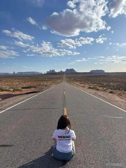Un capogruppo WeRoad è seduto su una lunga strada dritta che conduce a formazioni rocciose in un paesaggio desertico sotto un cielo blu.