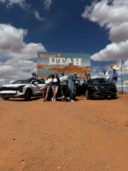 Un gruppo WeRoad posa per una foto con due auto davanti a un grande cartello 'Welcome to Utah' in un paesaggio desertico.