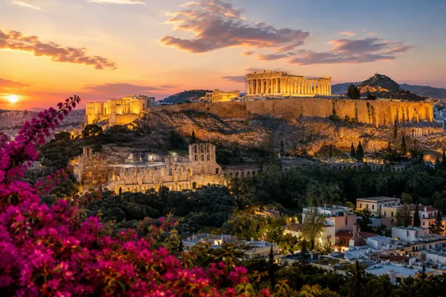 Antiche rovine su una grande collina rocciosa sono illuminate dal sole al tramonto, con una città sottostante e fiori rosa in primo piano.